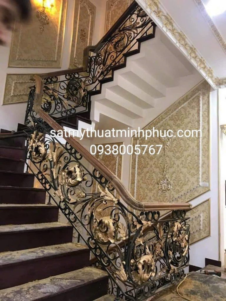Lan can cầu thang sắt mỹ thuật Minh Phúc thiết kế hoa văn xoắn ốc, khung sắt đen kèm chi tiết mạ vàng, tay vịn gỗ mahogany, nền tường trắng.