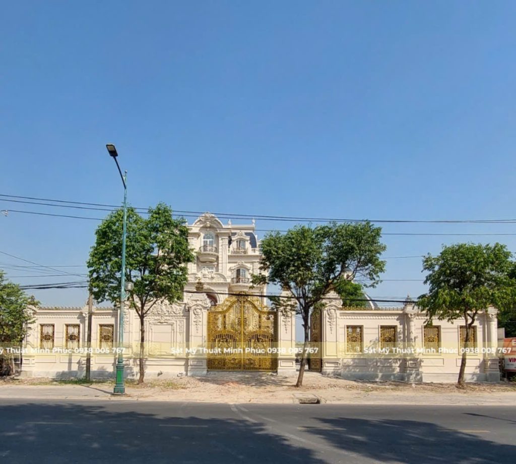 Lan can ban công sắt mỹ thuật màu đen – đồng, hoa văn lồng ghép họa tiết hoa lá cổ điển, lắp đặt trên tường ốp đá trắng, sân nền đá sỏi phía dưới.